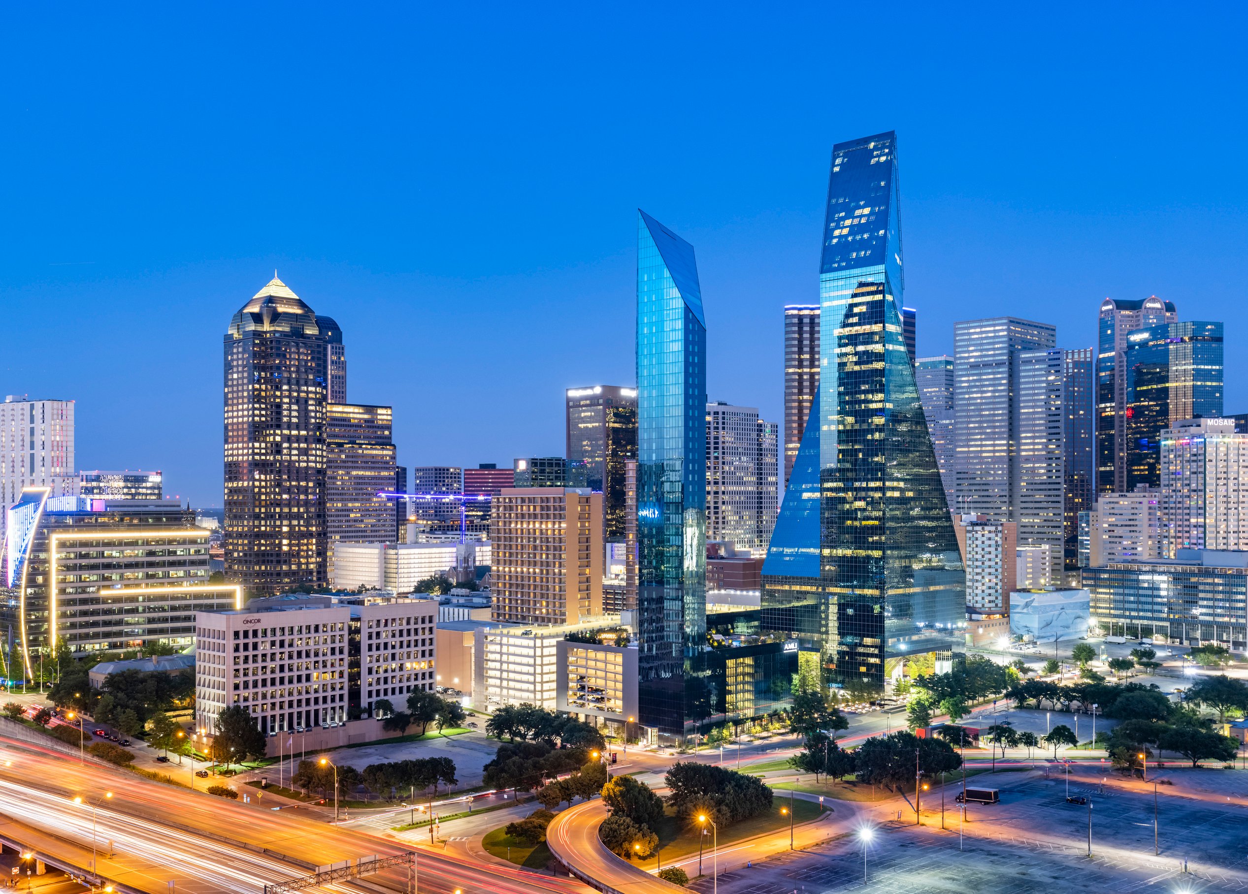 Two modern high rise apartments are featured in the center of a busy city at dusk.